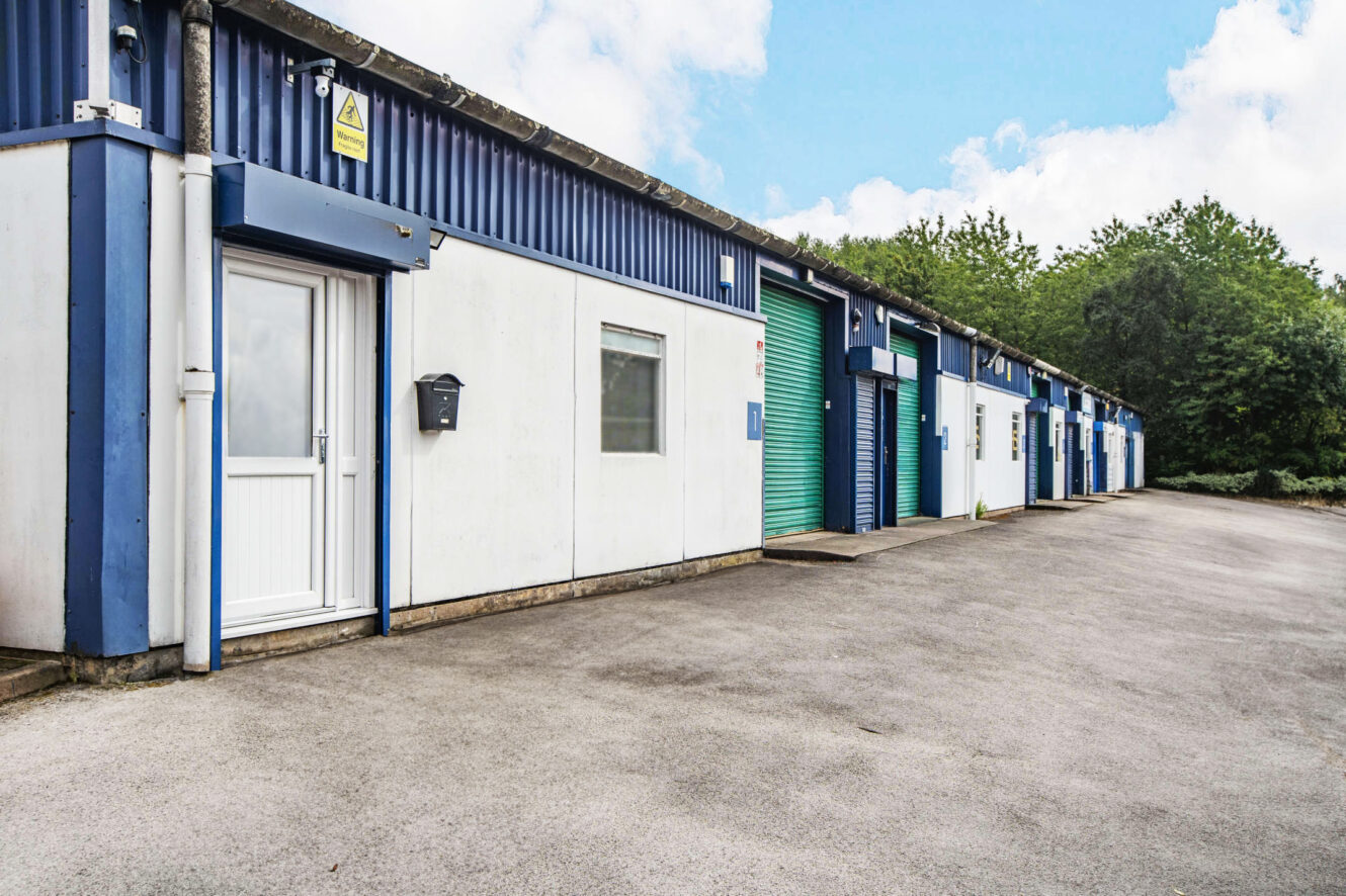 Row of industrial units with blue and white exteriors, closed doors, and green garage shutters, situated on a paved lot under a partly cloudy sky.