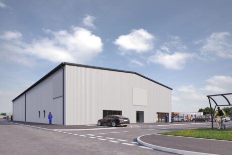 A large, light gray industrial warehouse with a black roof, surrounded by a parking lot with parked cars and a few people walking nearby under a partly cloudy sky.