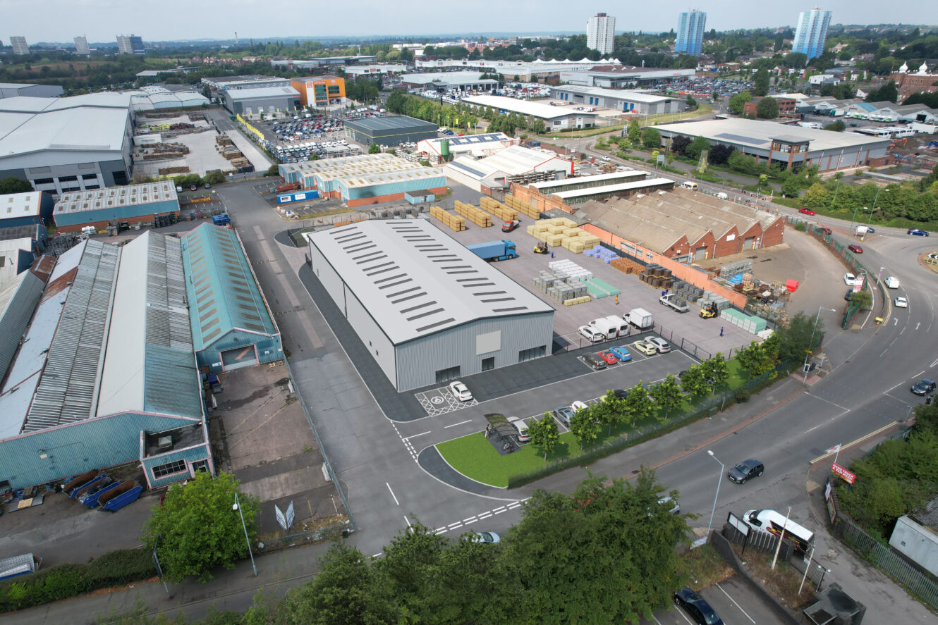 Aerial view of an industrial area with warehouses, storage yards, parked vehicles, and surrounding roads, with city buildings visible in the background.