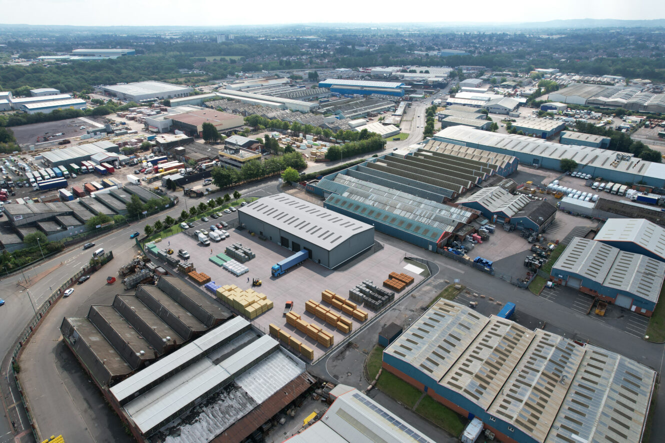 Aerial view of an industrial estate with multiple warehouses, factories, parked trucks, storage containers, and surrounding roads.