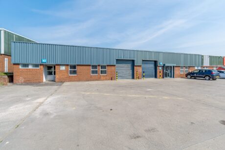 Single-story industrial warehouse with brick and metal exterior, three roller shutter doors, and two parked cars on a large concrete lot under a clear sky.