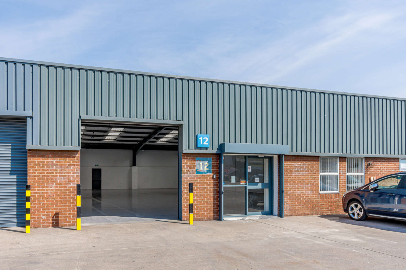 Exterior of an industrial warehouse unit with a brick and metal facade, labeled number 12, featuring a shutter door, glass entry, and a parked car partially visible on the right.