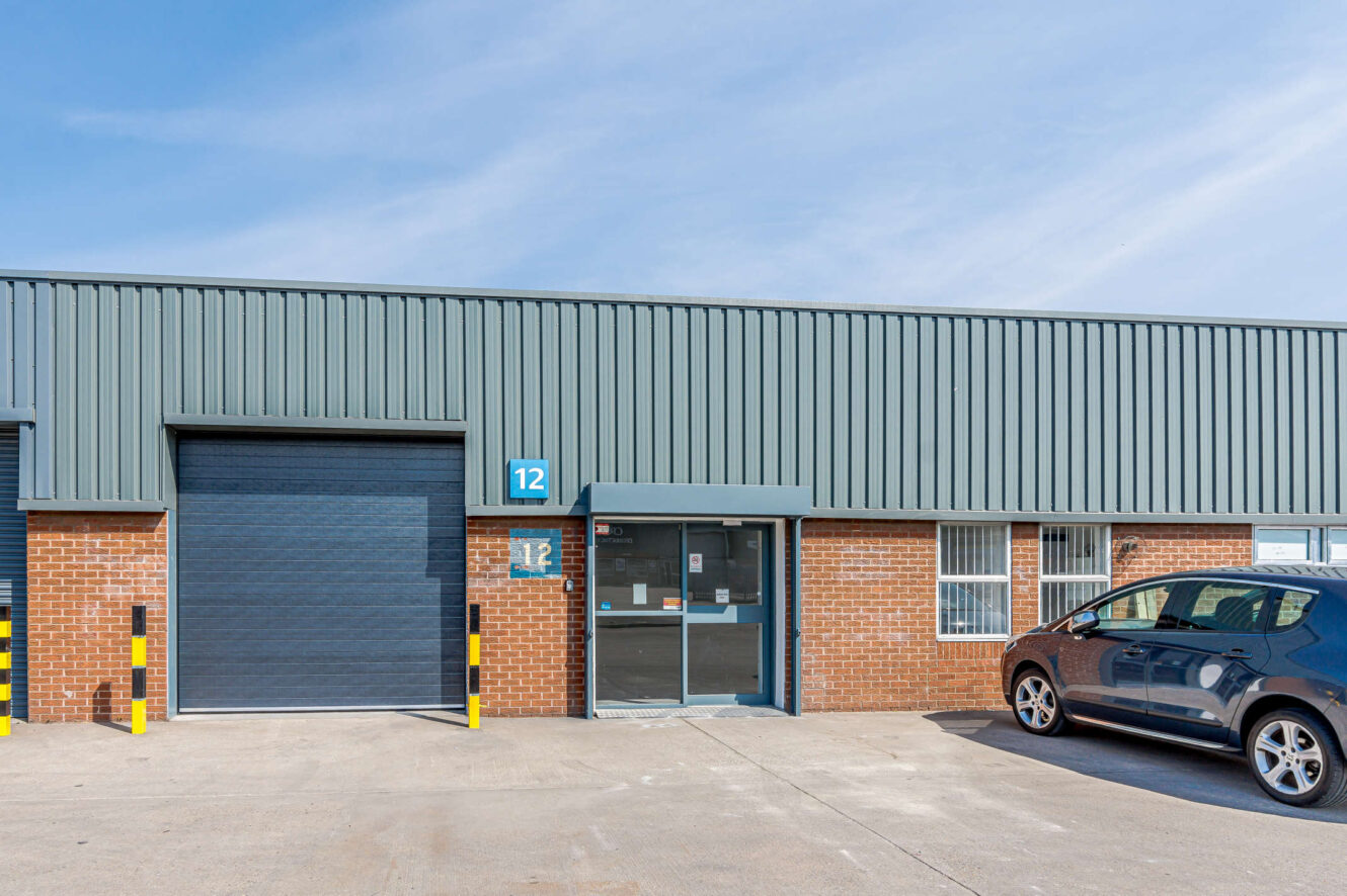Single-story industrial building with a grey roller door, glass entrance, unit number 12 sign, and a parked black car in front.