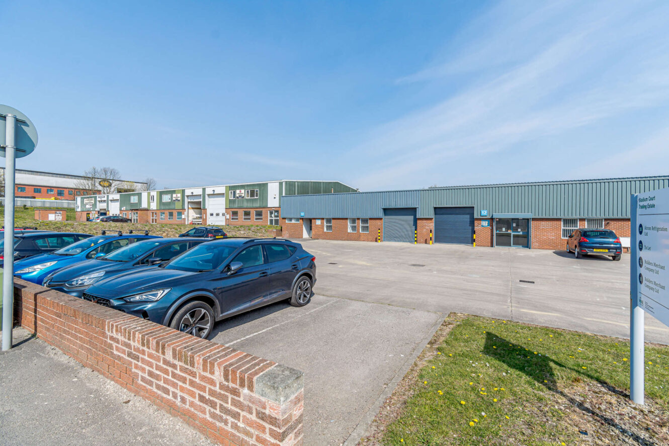 Several cars parked in front of a low industrial building with green and brick exterior on a clear day. A signpost and brick wall are visible in the foreground.