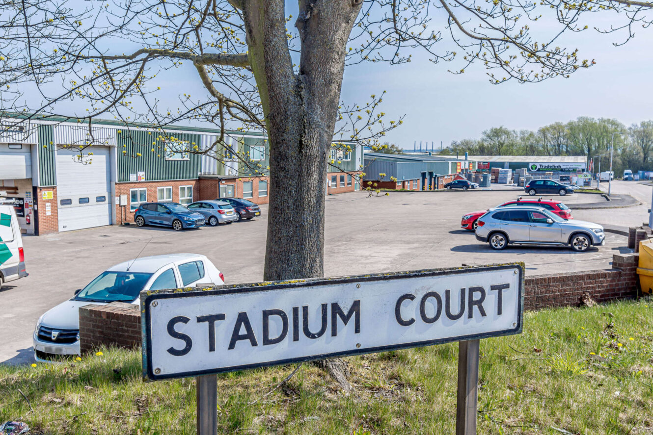 A tree stands near a Stadium Court sign in front of a parking lot and industrial buildings with several parked cars on a clear day.