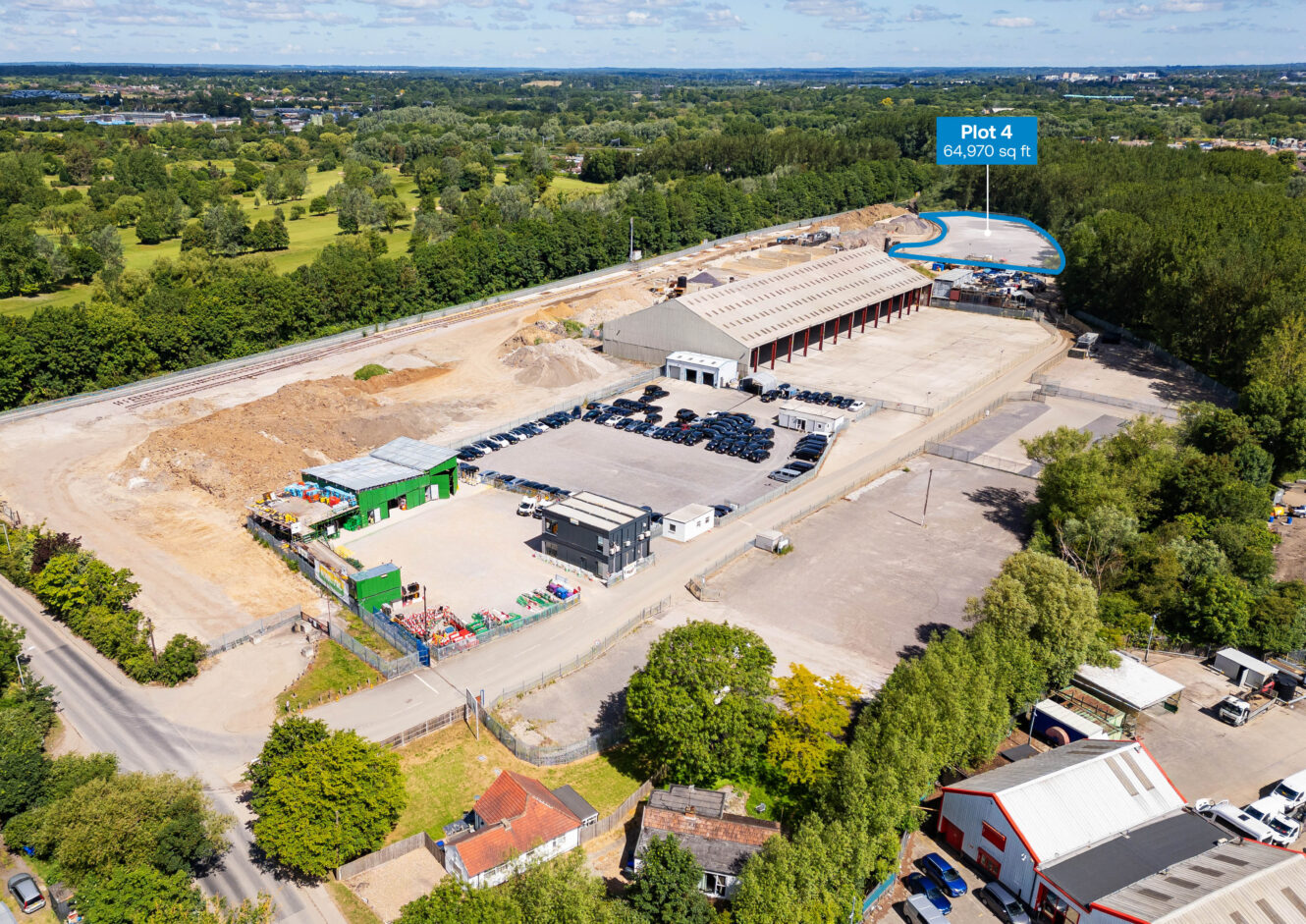 Aerial view of an industrial site with several buildings, parking areas, and a highlighted plot labeled “Plot 4 64,970 sq ft” in the upper right area, surrounded by trees.