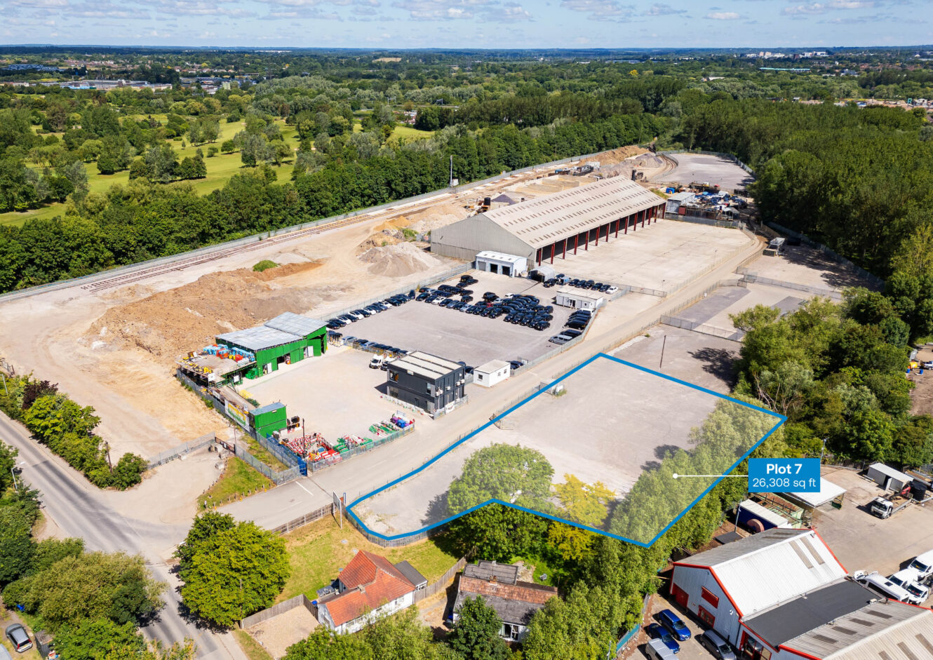 Aerial view of an industrial site with a marked vacant plot labeled Plot 7, 28,308 sq ft located near a road, surrounded by other buildings, greenery, and parked vehicles.