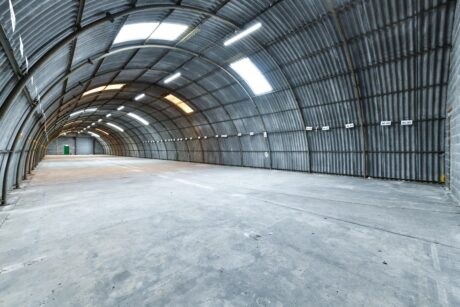 Wide view of an empty, arched industrial warehouse with corrugated metal walls and ceiling, concrete floor, fluorescent lights, and a green door in the distance.