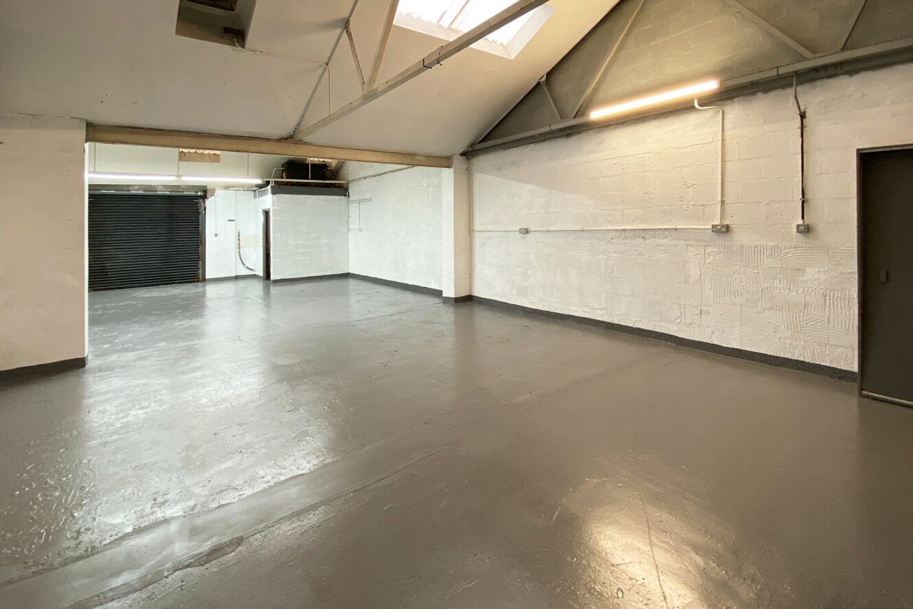 Empty industrial space with painted concrete floors, white brick walls, exposed beams, skylights, overhead lighting, and a black roller shutter door.