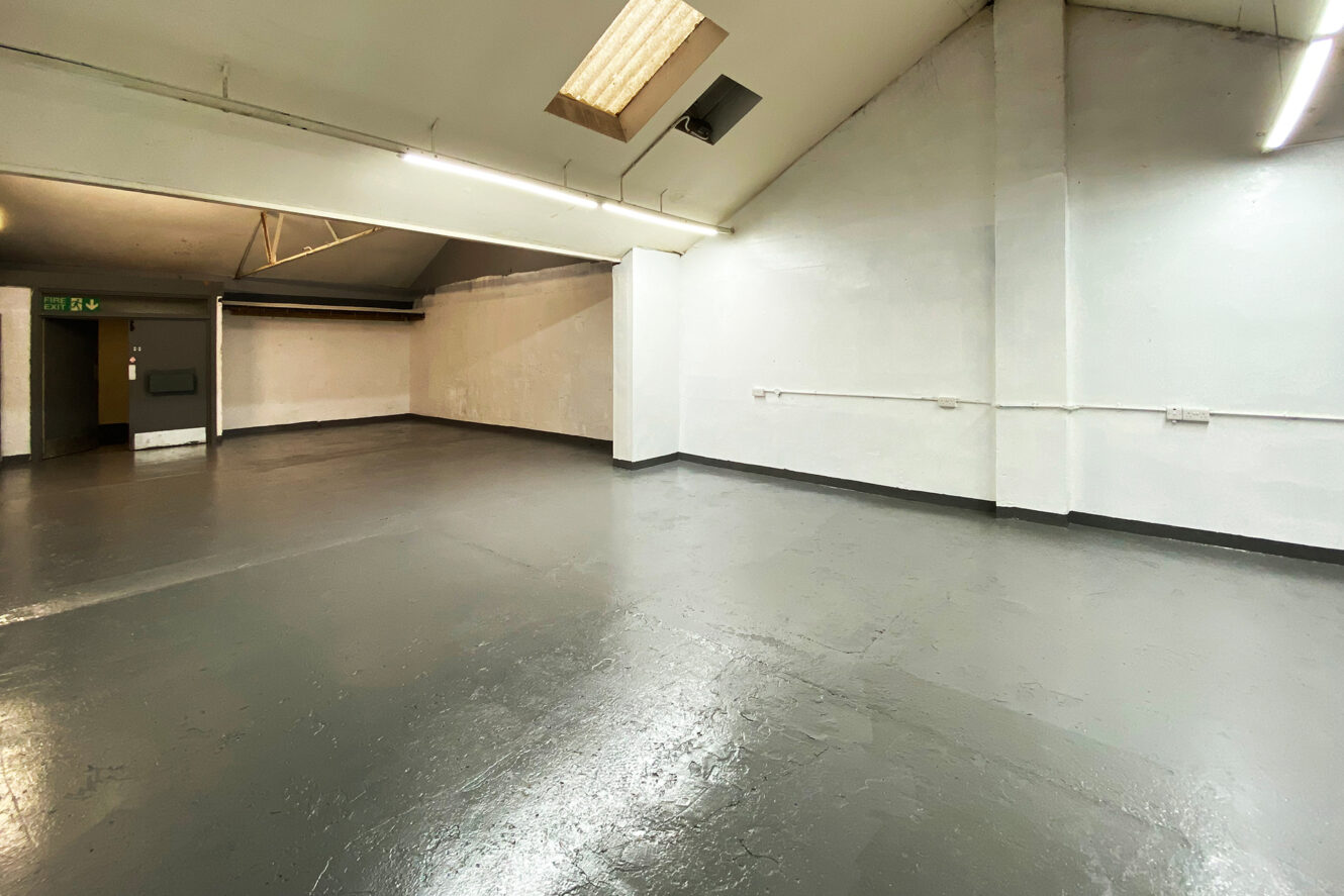 Empty, industrial-style room with smooth gray flooring, white painted walls, overhead fluorescent lighting, and exposed pipes along the walls.