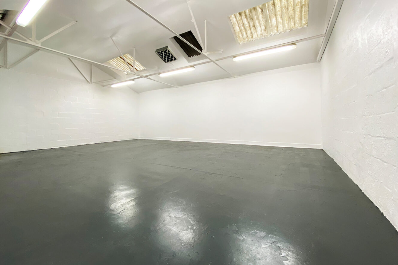Empty, white-walled industrial room with a grey floor, high ceiling, fluorescent lighting, and exposed beams. Some ceiling panels are damaged or missing.
