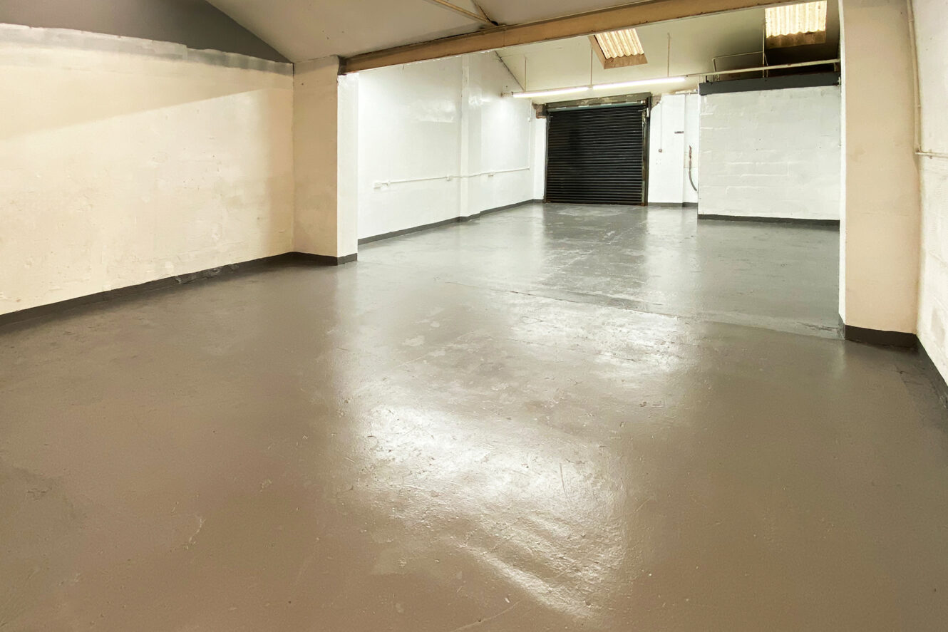 Empty industrial garage space with smooth concrete floor, white painted walls, overhead fluorescent lights, and a black roller shutter door at the far end.