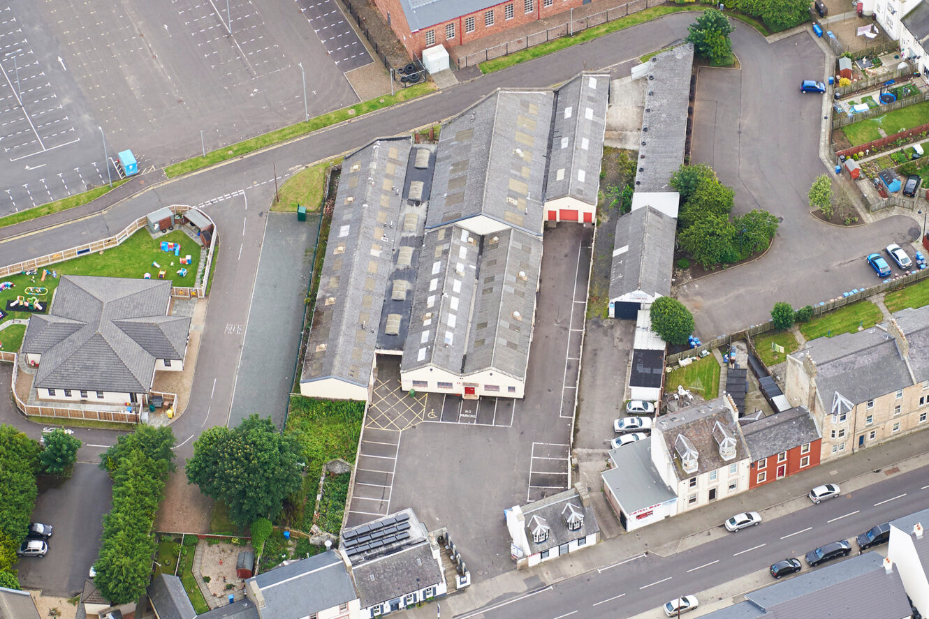 Aerial view of an industrial building complex surrounded by parking lots, roads, residential houses, and a small green play area.