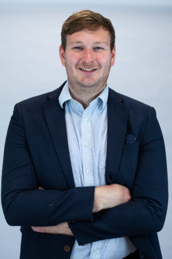 A man in a navy blazer and light blue shirt stands with arms crossed, smiling against a plain light background.