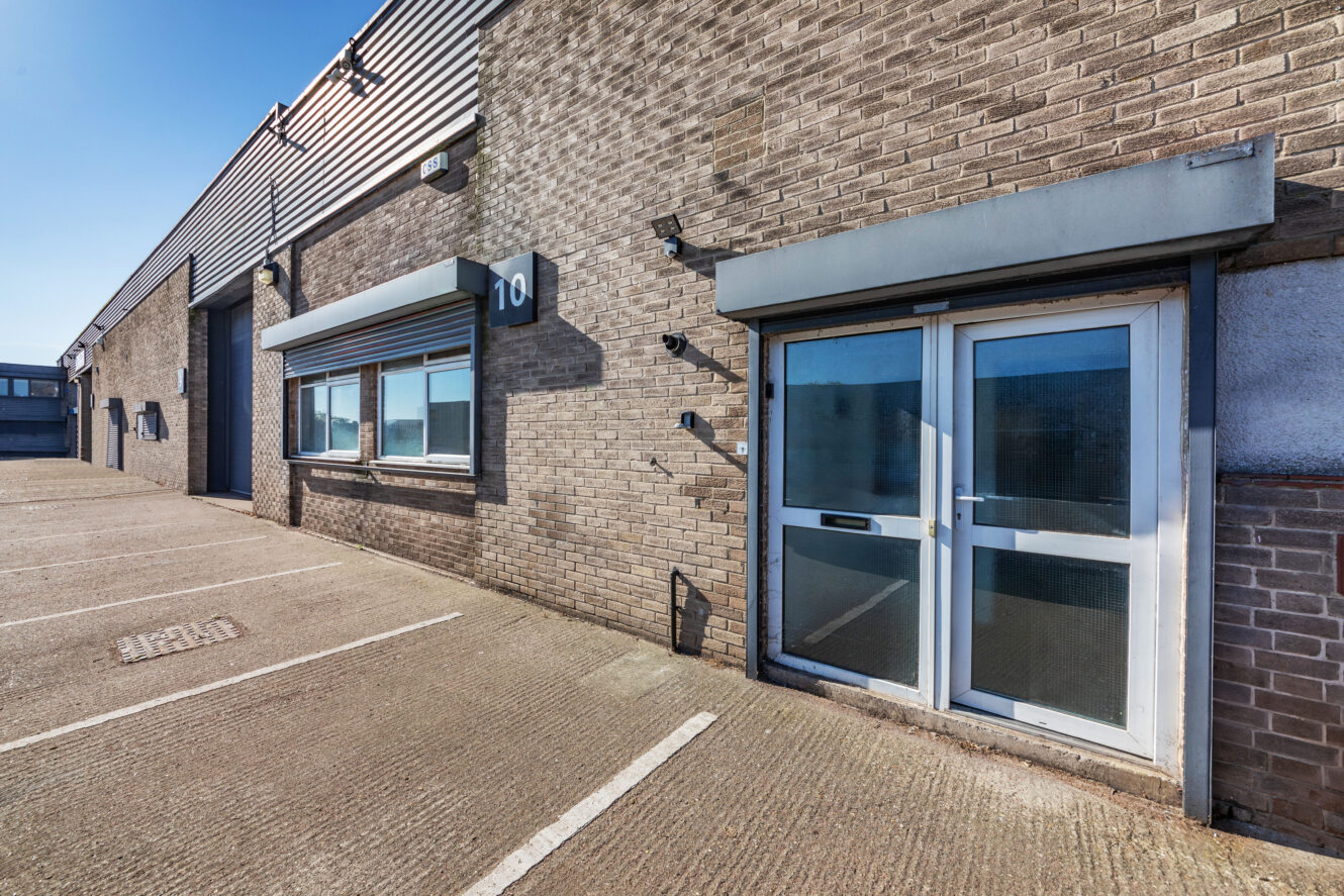 Exterior of an industrial warehouse unit with two metal doors, large windows, and numbered sign 10, next to an empty parking area under a clear sky.
