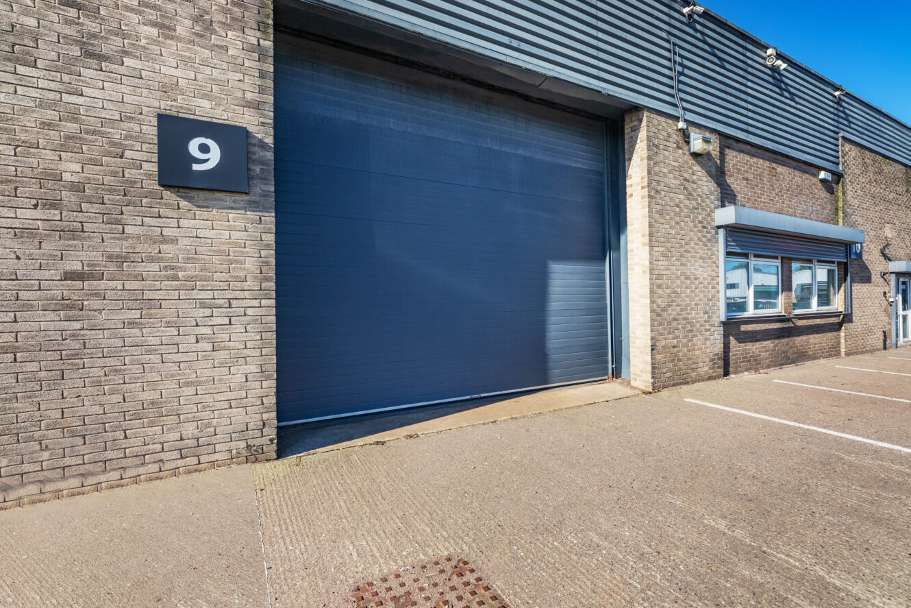 Exterior view of an industrial warehouse unit with a large closed gray shutter door and a sign displaying the number 9 on the brick wall.