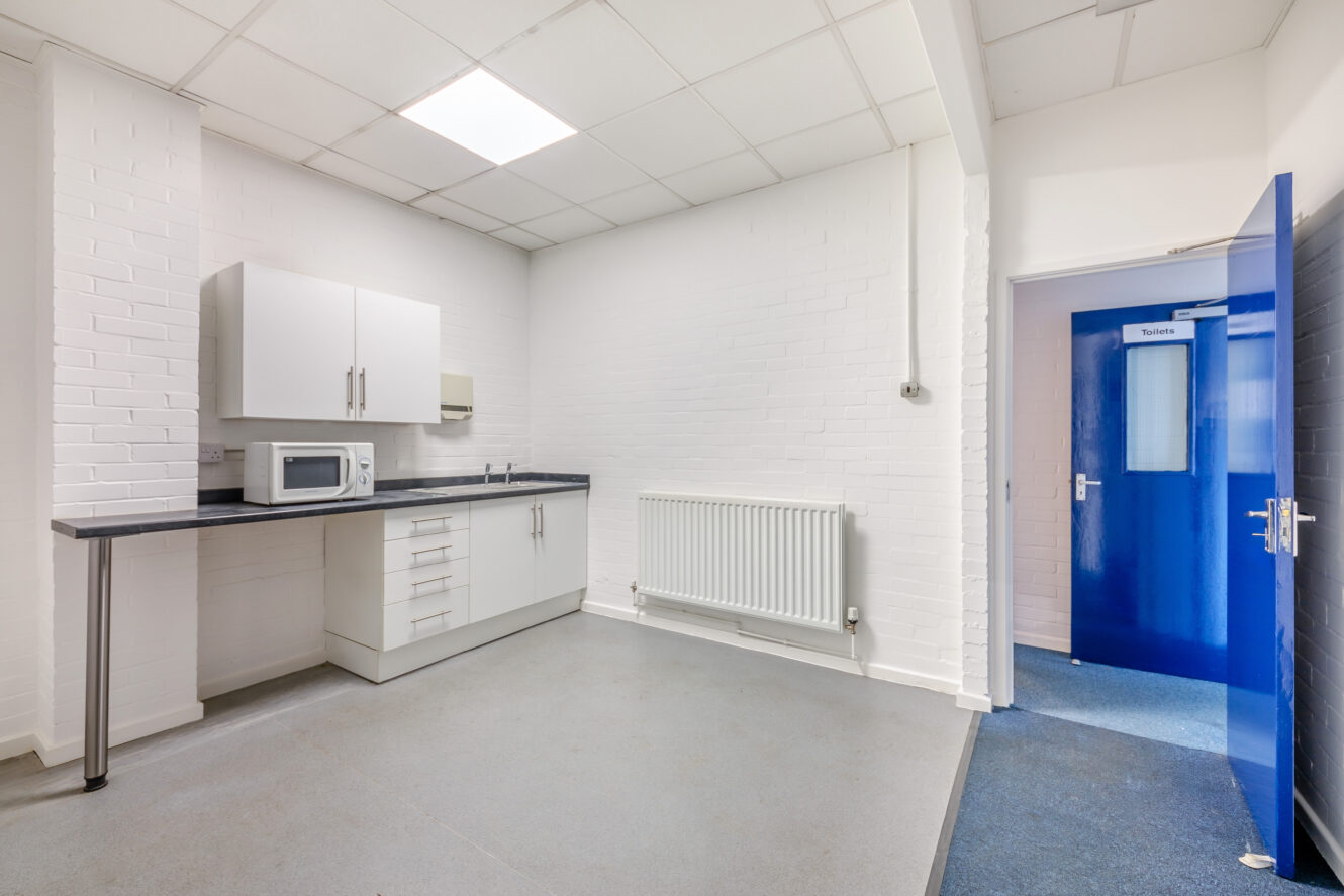 Small kitchenette with white cabinets, a microwave, a sink, and a radiator, next to a blue door labeled Toilets in a clean, bright room with white brick walls.