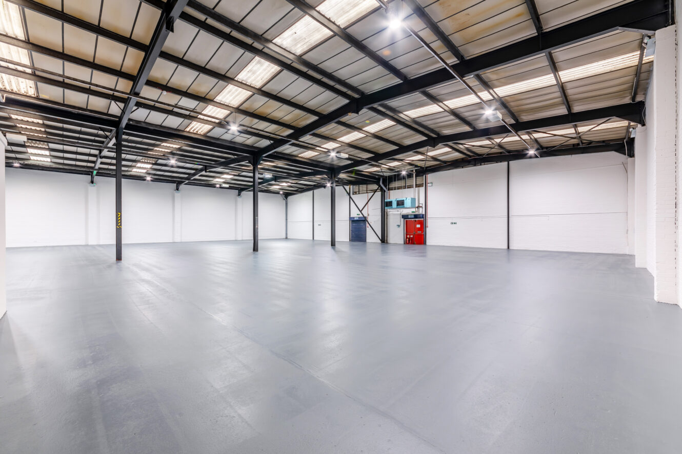 Large, empty industrial warehouse with polished concrete floors, high ceilings, metal beams, white walls, and overhead fluorescent lighting.