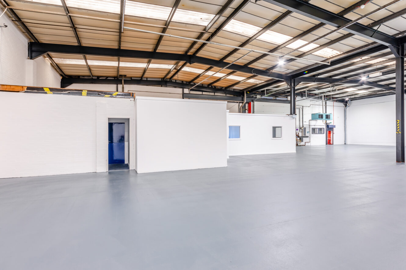Interior of a large empty warehouse with a small white office structure, gray flooring, high ceilings, and visible overhead lighting.
