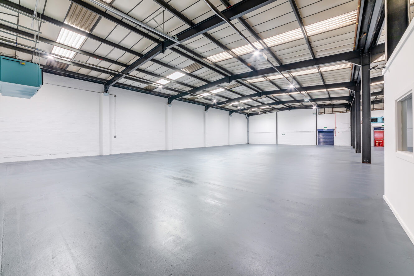 Large, empty industrial warehouse with white walls, high ceiling, exposed beams, and polished concrete floor. Bright lighting and a few windows provide natural light.