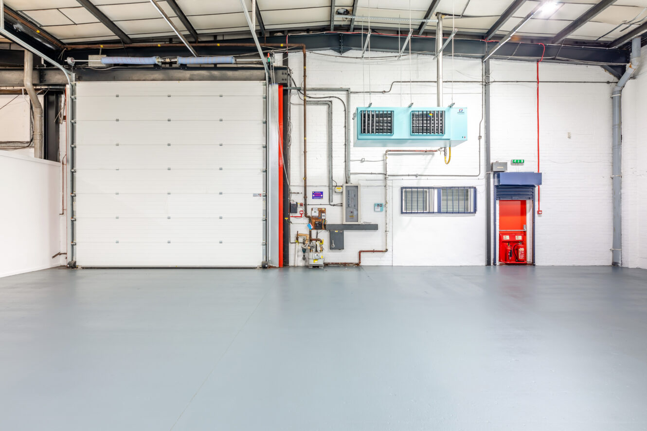 Empty industrial warehouse interior with a large roller shutter door, utility panels, and emergency fire equipment near a red exit door. The floor is clean and the space is well-lit.