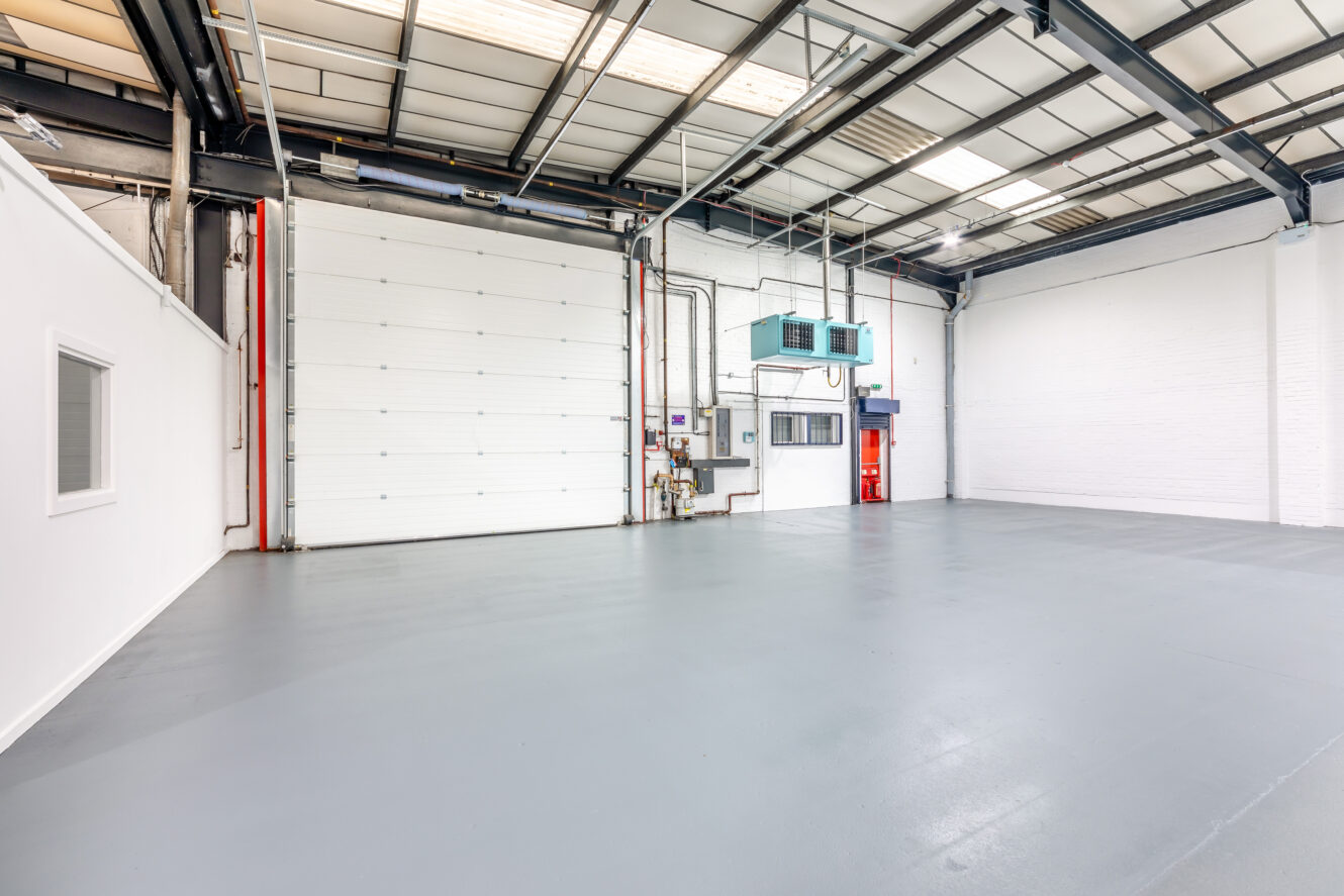 An empty industrial warehouse with a large roller door, white walls, grey flooring, overhead lighting, and exposed pipes and cables.
