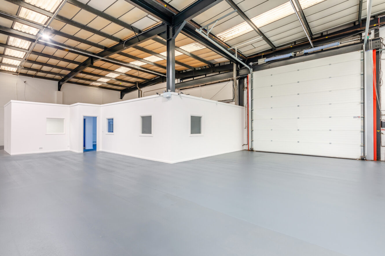 Interior of an empty warehouse with white office partition, grey epoxy floor, high ceiling, and large rolling garage door.