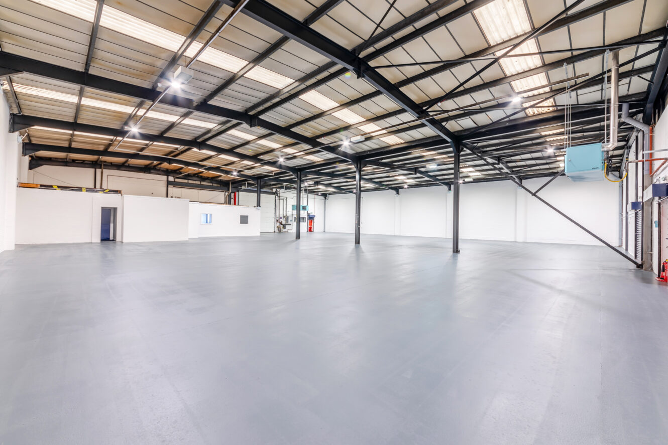 Spacious, empty industrial warehouse with high ceilings, exposed steel beams, white walls, and large windows for overhead natural lighting.