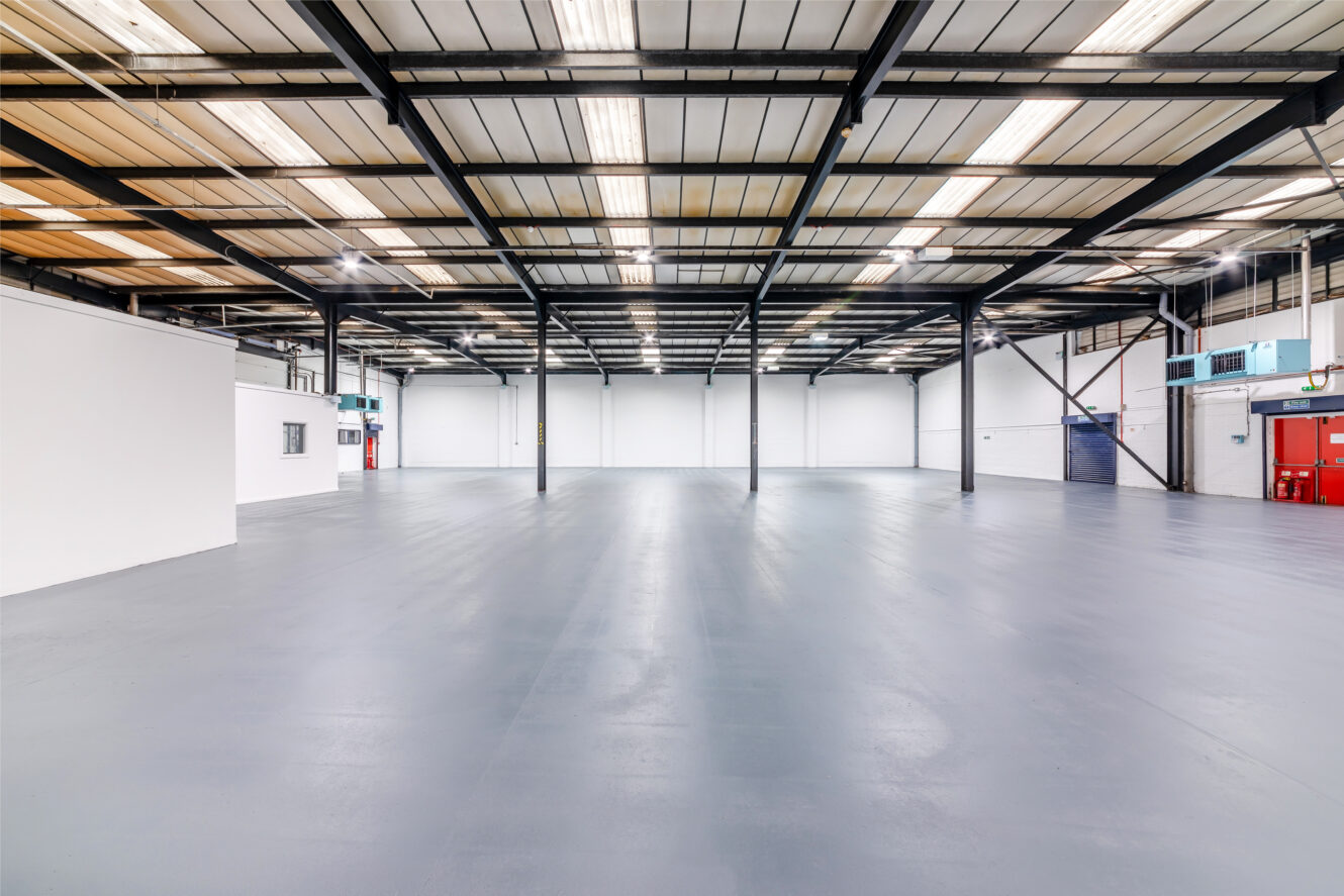 Spacious, empty industrial warehouse with a high ceiling, exposed beams, white walls, and polished concrete floor, illuminated by overhead fluorescent lights.