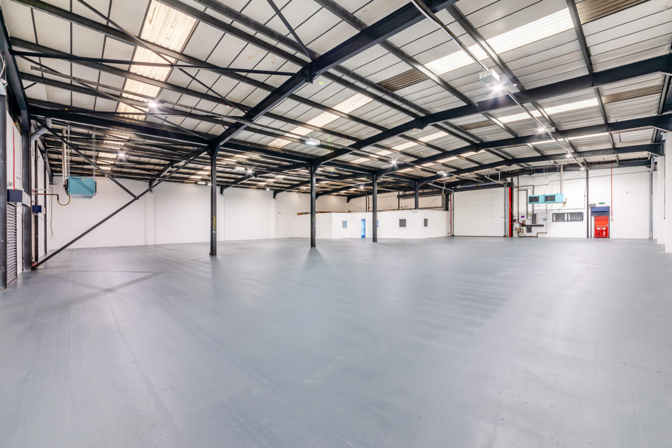 Interior view of an empty industrial warehouse with a clean, gray floor, white walls, steel beams, and overhead lighting. Large loading doors and windows are visible at the far end.