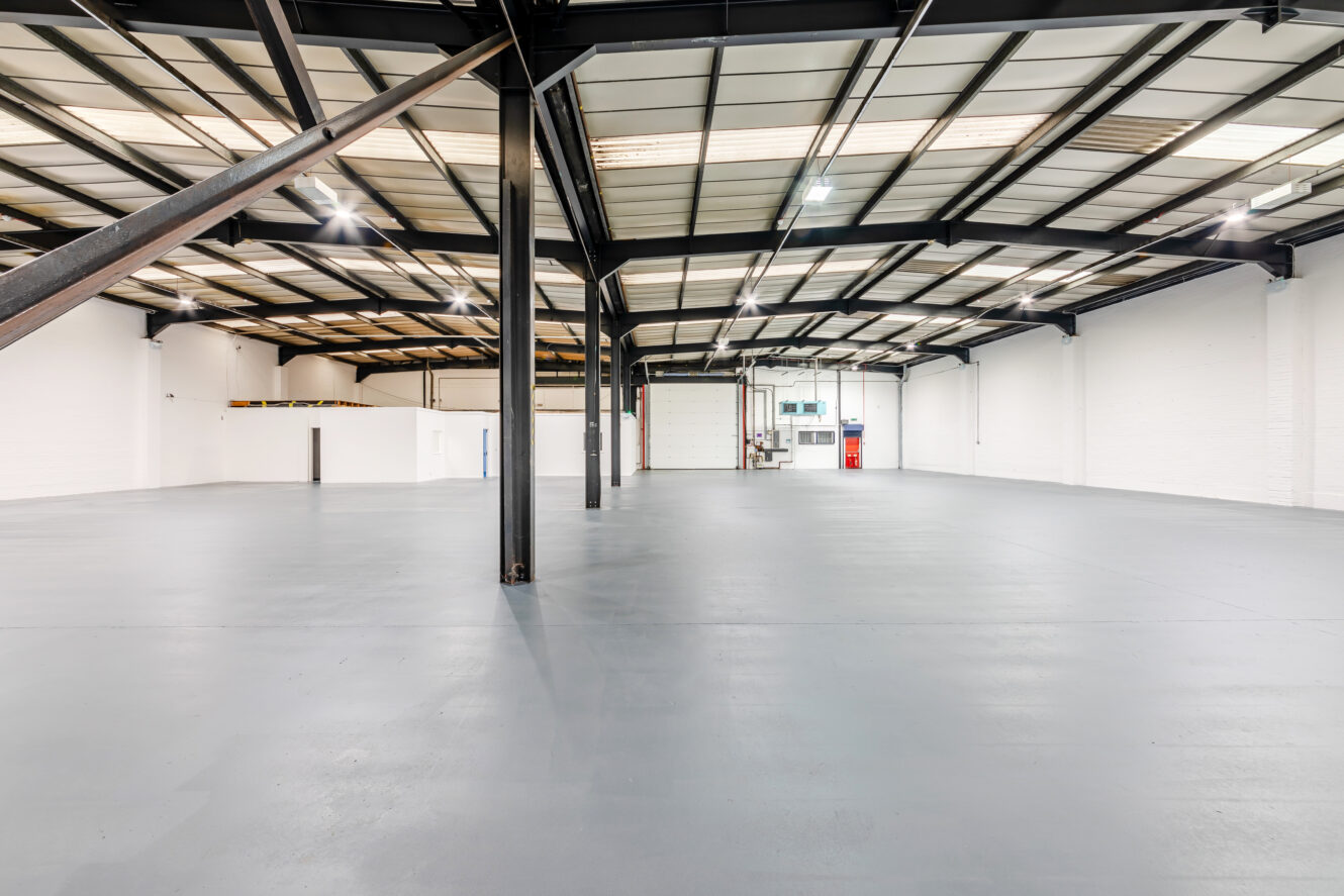 Spacious, empty industrial warehouse with high ceilings, exposed beams, white walls, and a smooth concrete floor.