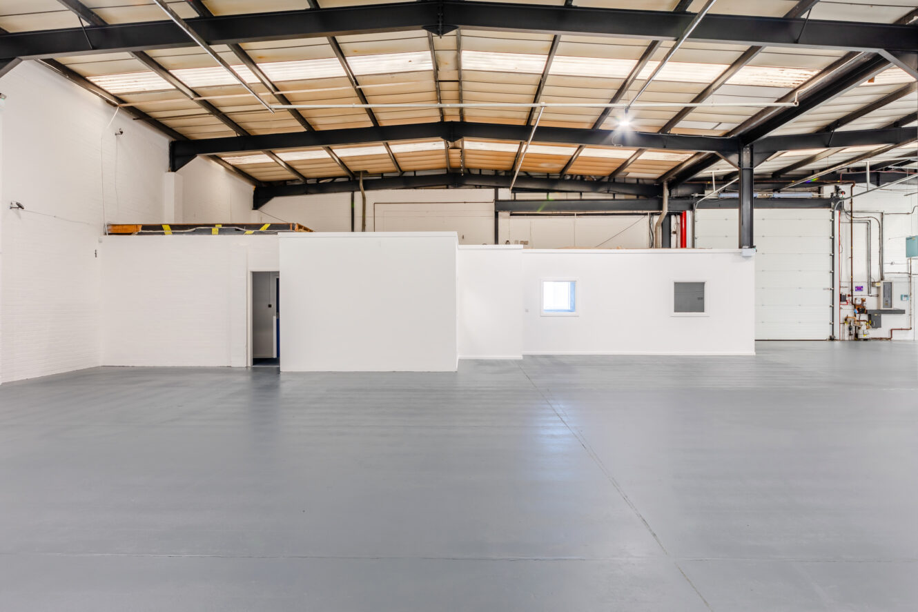 Large empty warehouse interior with gray floor, white walls, exposed beams, and a small enclosed office area with windows and doors.