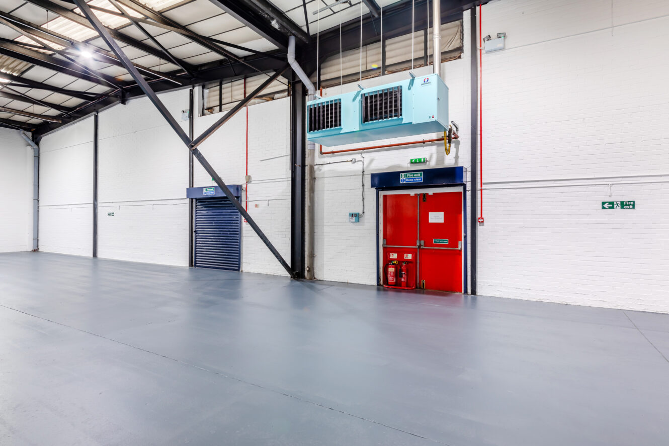 Empty industrial warehouse interior with polished concrete floor, white brick walls, exposed metal beams, red emergency exit door, blue roller shutter, and suspended air conditioning units.