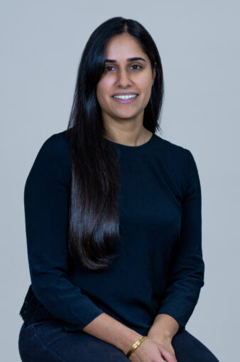A woman with long dark hair, wearing a black long-sleeve top and dark pants, sits and smiles at the camera against a plain light background.