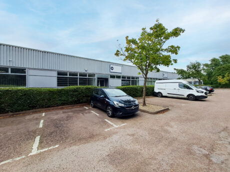 A small parking lot with two cars parked in front of a low, modern industrial building on a cloudy day.