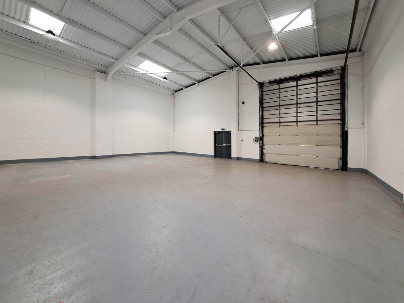 Empty industrial warehouse with high ceilings, concrete floor, white walls, skylights, a large roller door, and a small black exit door.