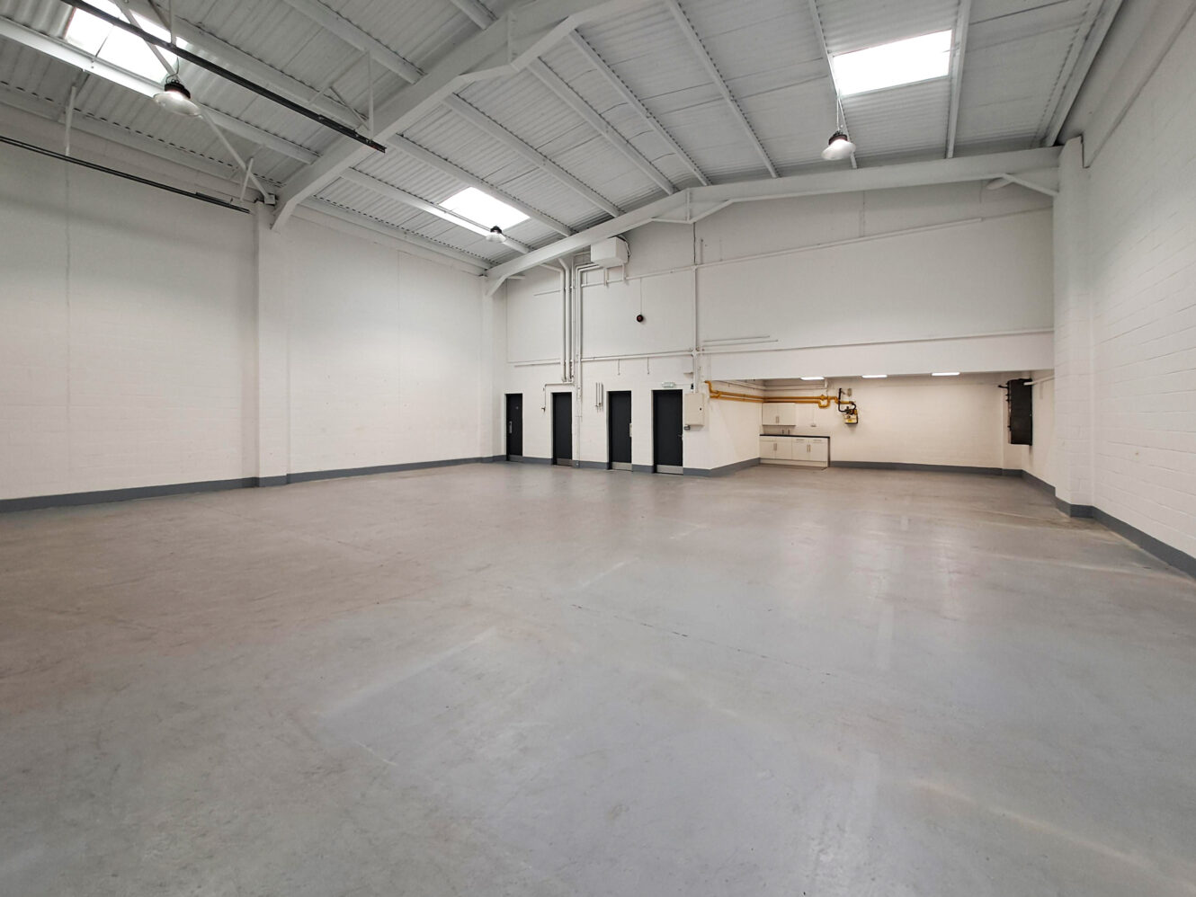 Spacious empty warehouse with high ceilings, white walls, concrete floor, and several black doors along the back wall under fluorescent lighting.