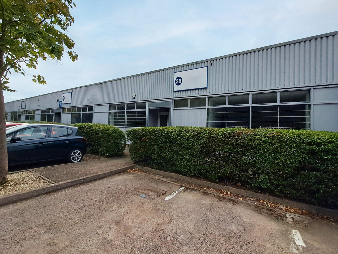 A grey industrial building with large windows, marked with the number 36, is surrounded by hedges and a parked car in front.