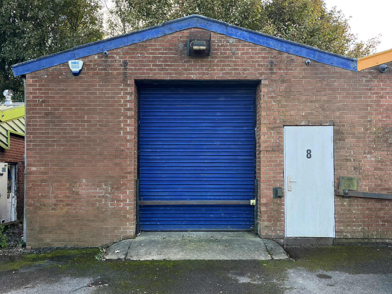 A brick building with a blue roll-up garage door and a white door labeled 8 on the right side. Trees and part of a yellow structure are visible in the background.
