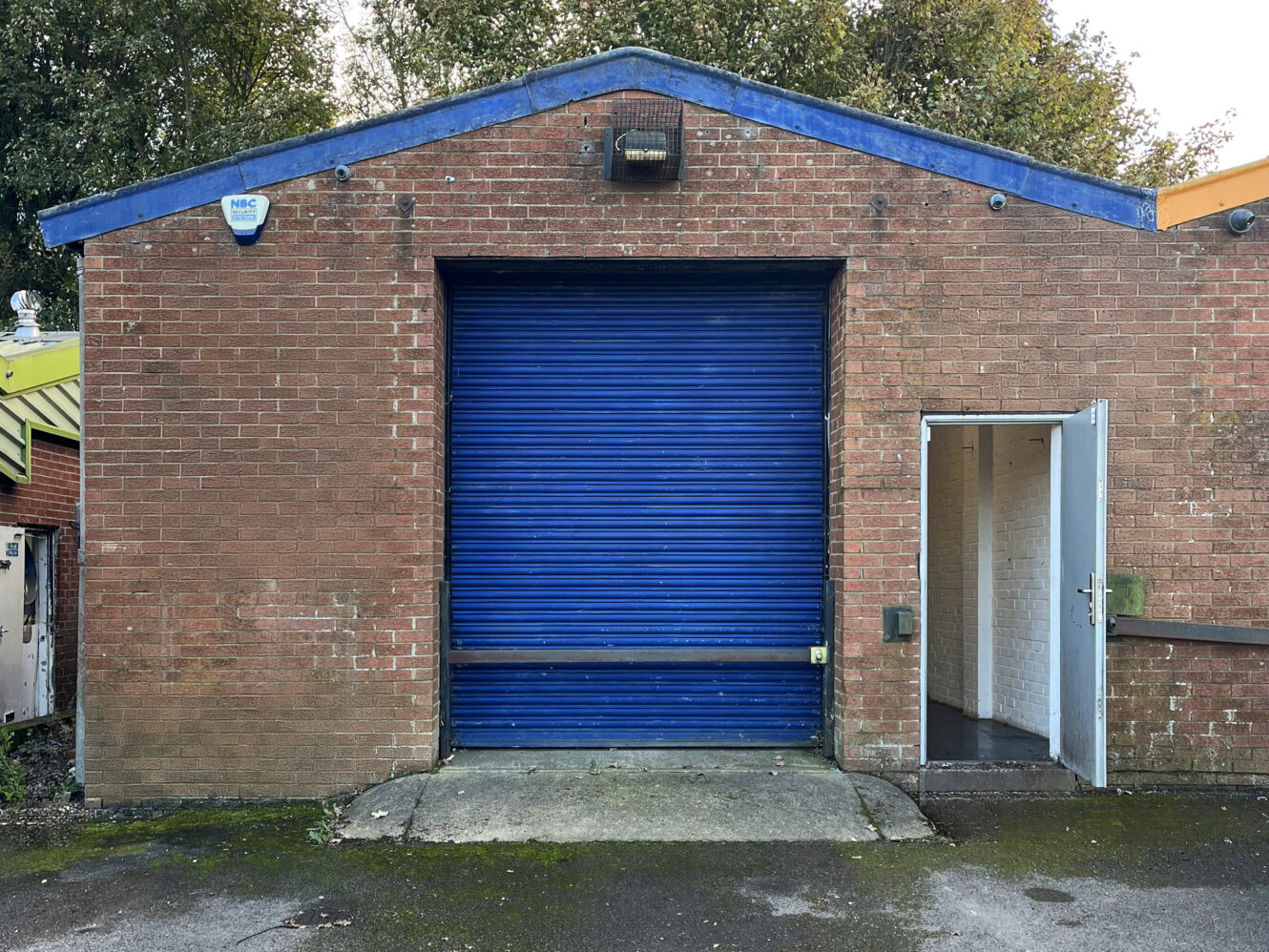 A brick industrial building with a closed blue roller shutter door and an open white side door leading inside.