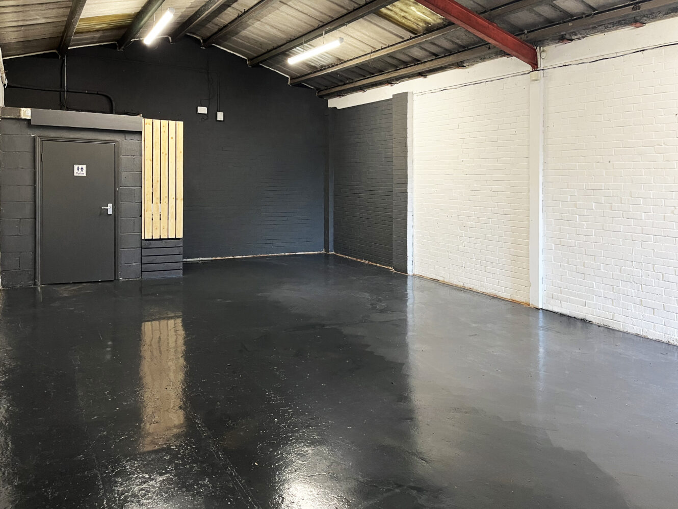 An empty industrial room with black and white painted brick walls, a polished black floor, exposed ceiling beams, and a door marked “11” on the left.