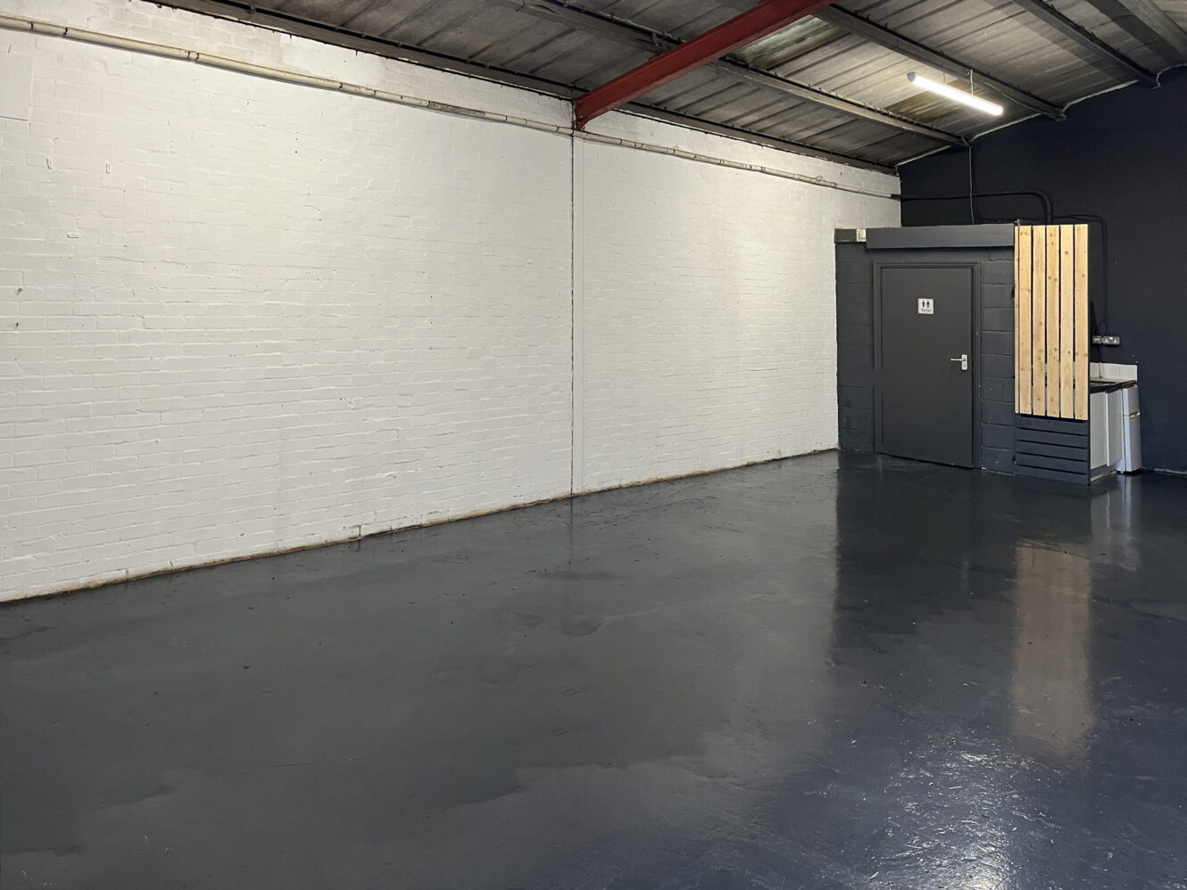Empty industrial room with white brick walls, a grey painted floor, exposed metal ceiling, and a closed grey door in the corner.