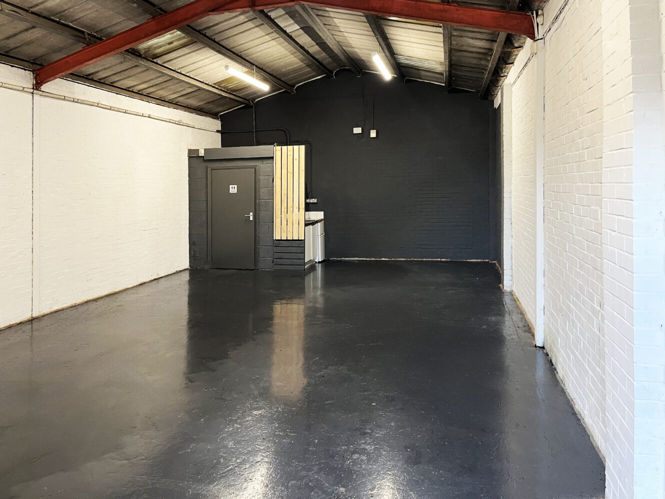 Industrial-style empty room with a concrete floor, white brick walls, a small kitchenette, and a gray door under a pitched ceiling with exposed beams.