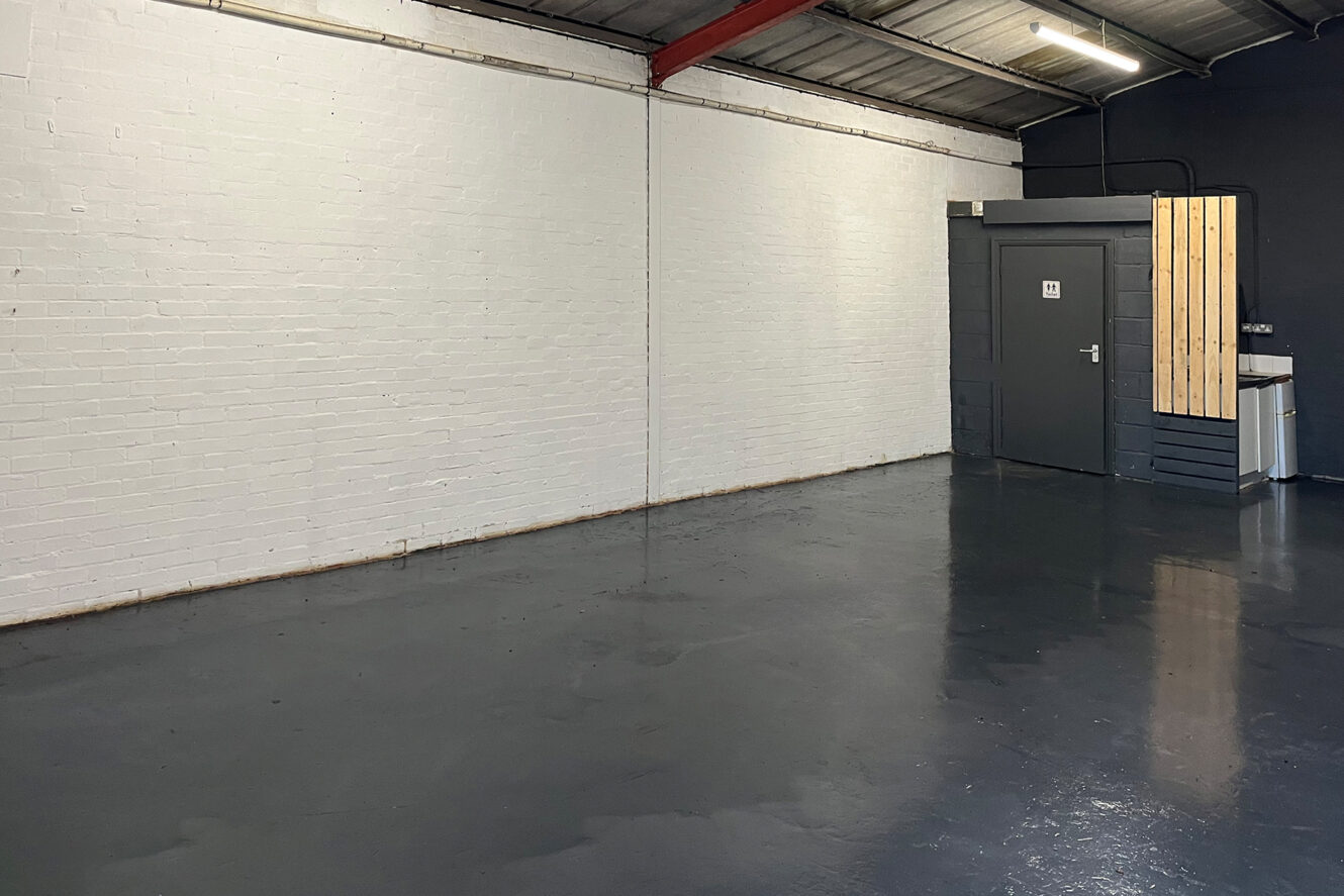 Empty industrial room with white brick walls, gray polished floor, exposed ceiling beams, and a door labeled 11 near a small kitchenette in the corner.