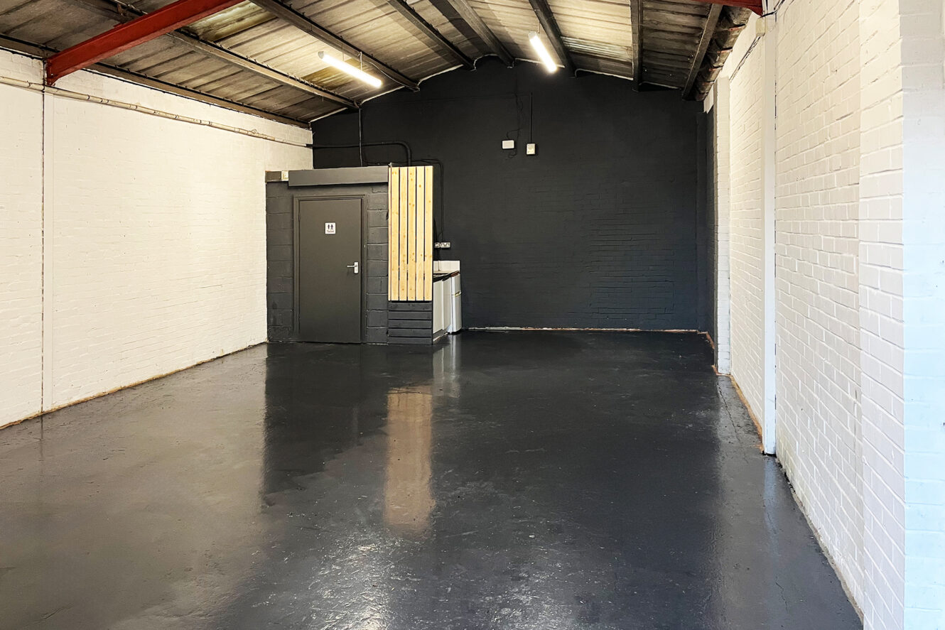 Empty industrial-style room with black polished floor, white brick walls, exposed ceiling beams, and a small enclosed area with a door and countertop at the back.