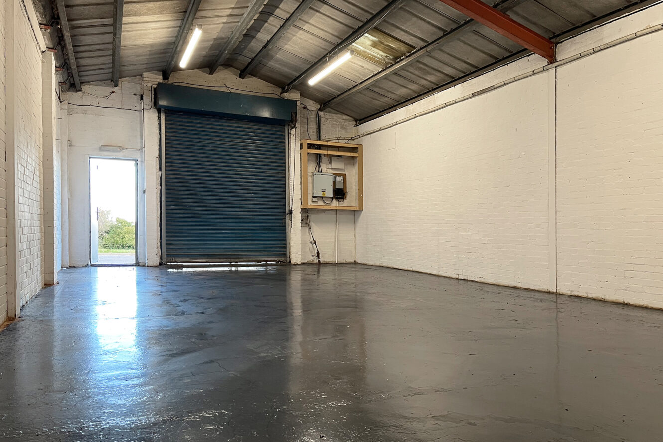 An empty industrial warehouse with white brick walls, a polished concrete floor, a blue roller shutter door, and exposed metal ceiling beams.