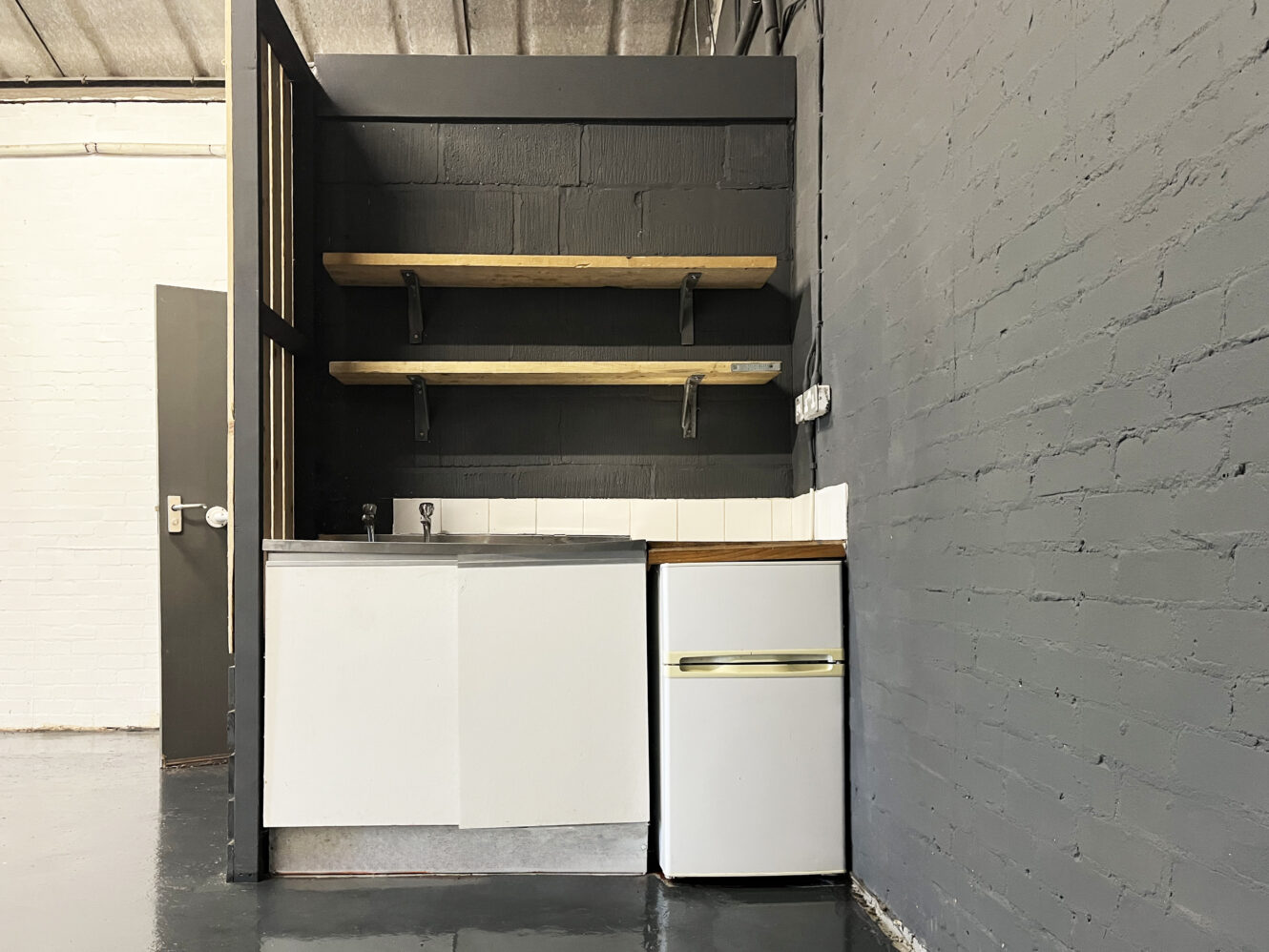 Small kitchenette with a white sink unit, compact fridge, tile backsplash, two wooden shelves, and gray painted brick walls.