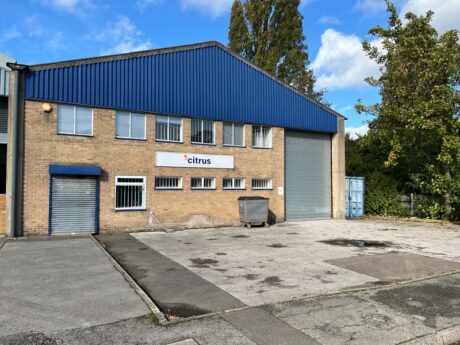 A brick industrial building with a blue roof, a sign that reads citrus, a large roller door, a metal dumpster, and a shipping container outside.