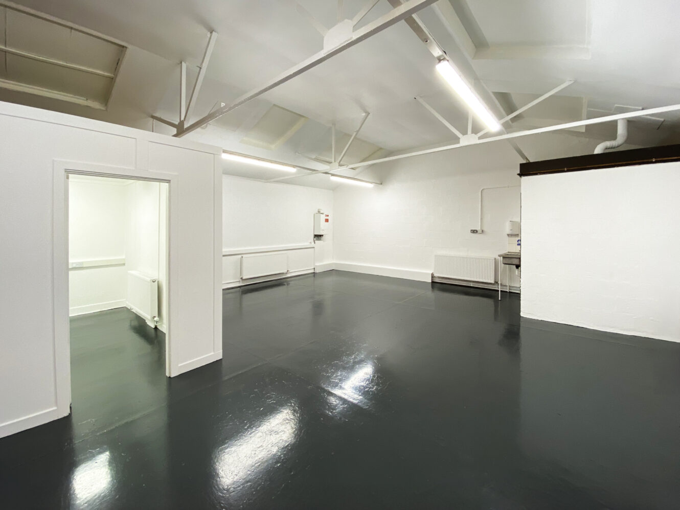 An empty, clean room with white walls, dark shiny floors, exposed beams, fluorescent lighting, and an adjacent small enclosed space.