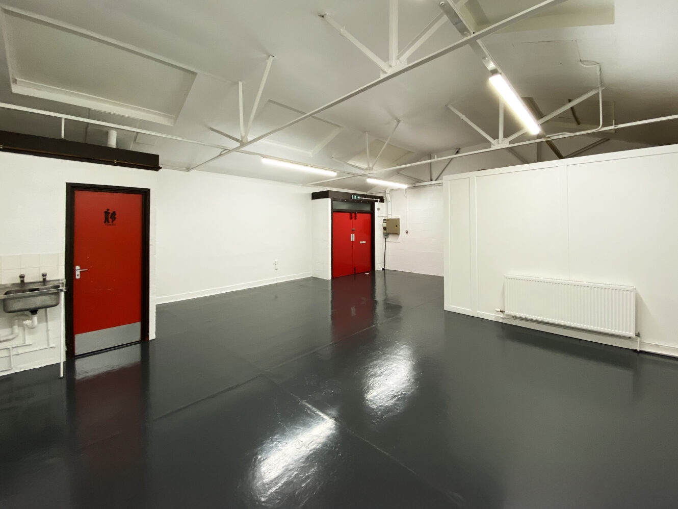 An empty industrial room with glossy dark floors, white walls, a red door, double red doors, a radiator, and a wall-mounted sink.